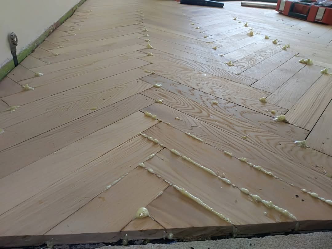 Parkiet w jodełkę, świeżo po klejeniu, z widocznymi punktami kleju. Ujęcie z niskiego kąta, ukazujące detale drewnianej podłogi i narzędzia w tle.