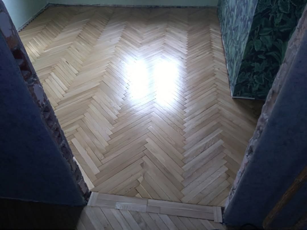 Jasny parkiet ułożony w jodełkę, świeżo po renowacji, z widocznym fragmentem ściany w ciemne, roślinne wzory. Widok z góry, z fragmentami futryn po bokach.