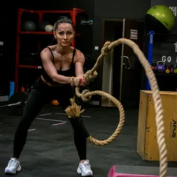 Kobieta w czarnym stroju sportowym ćwiczy z grubą liną w siłowni. W tle sprzęt do ćwiczeń i drabinka.