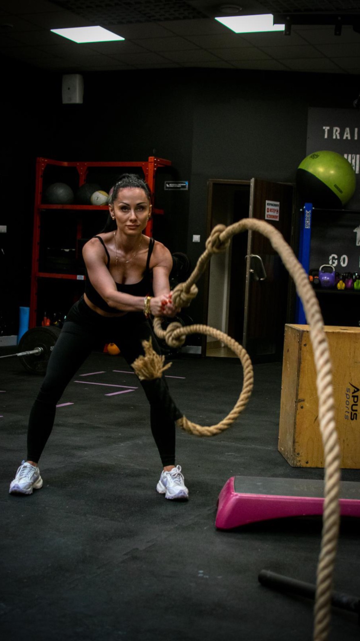 Kobieta w czarnym stroju sportowym ćwiczy z grubą liną w siłowni. W tle sprzęt do ćwiczeń i drabinka.