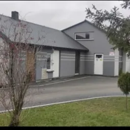 Budynek warsztatu samochodowego z logo 'Dobry Warsztat' na szczycie. Szary budynek z szarą kostką brukową i zielonym trawnikiem. Samochody zaparkowane w tle.