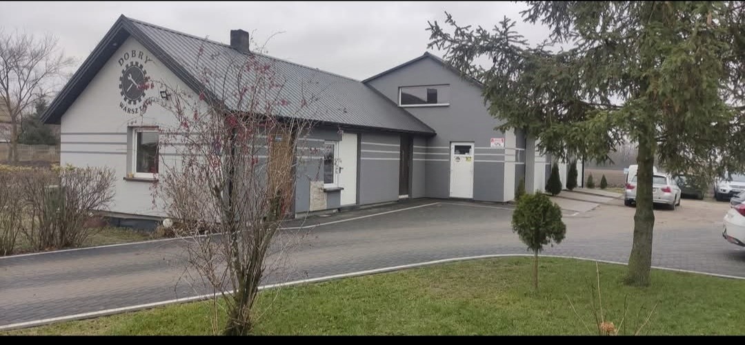 Budynek warsztatu samochodowego z logo 'Dobry Warsztat' na szczycie. Szary budynek z szarą kostką brukową i zielonym trawnikiem. Samochody zaparkowane w tle.