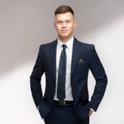 Młody mężczyzna w granatowym garniturze, biała koszula i wzorzysty krawat, z rękami w kieszeniach, prezentuje profesjonalny wizerunek na jasnym tle.