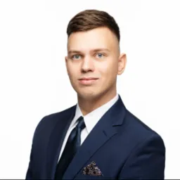 Portret młodego mężczyzny w ciemnym garniturze na jasnym tle. Widoczna poszetka w kieszonce, ciemny krawat i jasna koszula. Uśmiech na twarzy, profesjonalny wygląd.