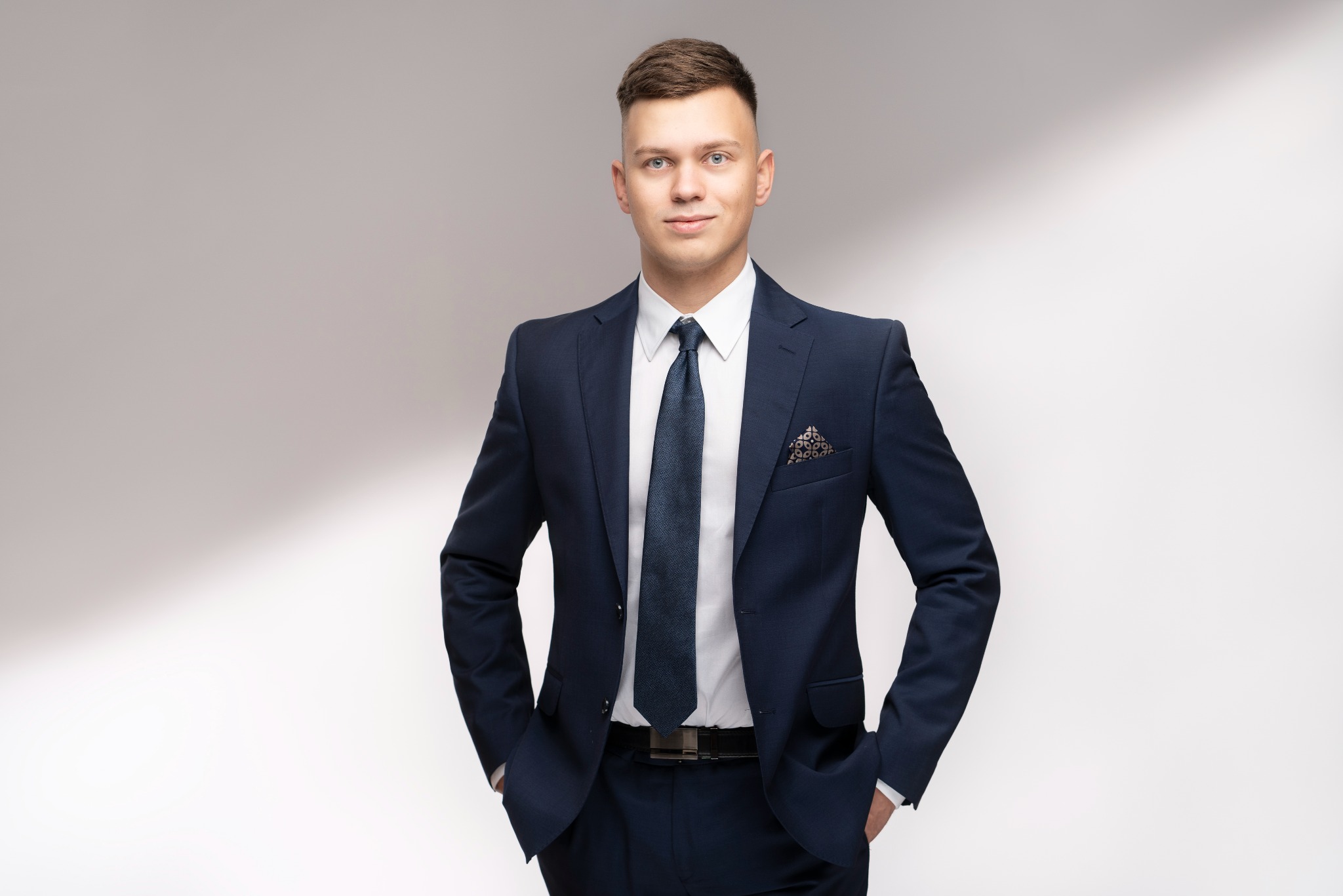 Młody mężczyzna w granatowym garniturze, biała koszula i wzorzysty krawat, z rękami w kieszeniach, prezentuje profesjonalny wizerunek na jasnym tle.