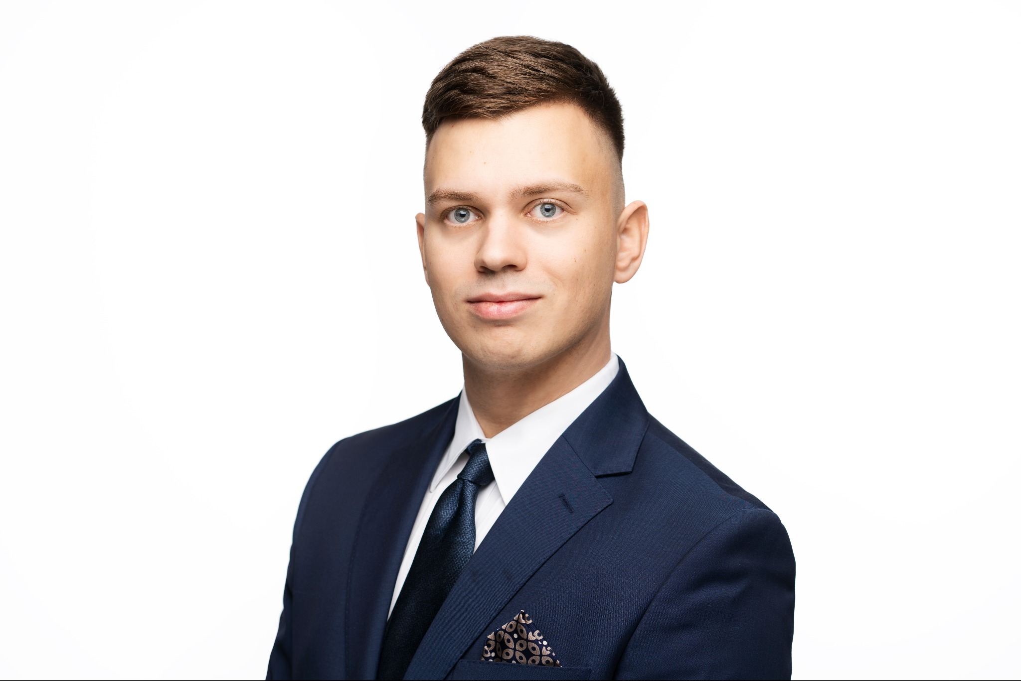 Portret młodego mężczyzny w ciemnym garniturze na jasnym tle. Widoczna poszetka w kieszonce, ciemny krawat i jasna koszula. Uśmiech na twarzy, profesjonalny wygląd.