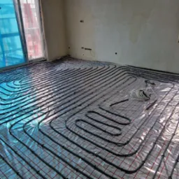 Instalacja ogrzewania podłogowego w pokoju z oknami, rury ułożone na folii z siatką, widoczne przyłącza do ściany, surowy stan wykończenia.