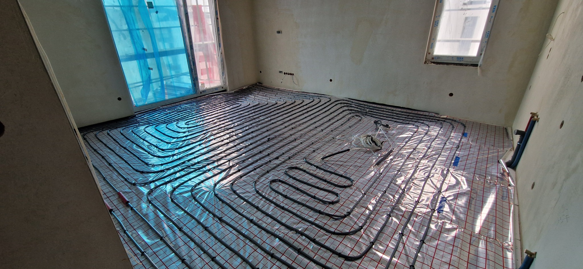 Instalacja ogrzewania podłogowego w pokoju z oknami, rury ułożone na folii z siatką, widoczne przyłącza do ściany, surowy stan wykończenia.