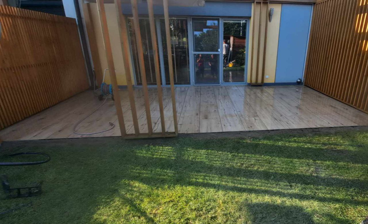 Nowoczesny taras z jasnej kostki imitującej drewno, przylegający do budynku z dużymi oknami i drewnianą ażurową ścianą. Widoczny trawnik i fragment narzędzi.