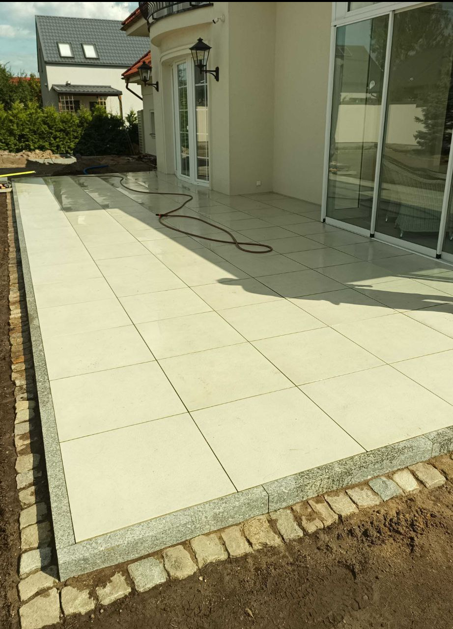 Wyłożony jasnymi płytami taras obramowany kostką granitową, z widocznym wężem ogrodowym i fragmentem domu z oknami i drzwiami balkonowymi.