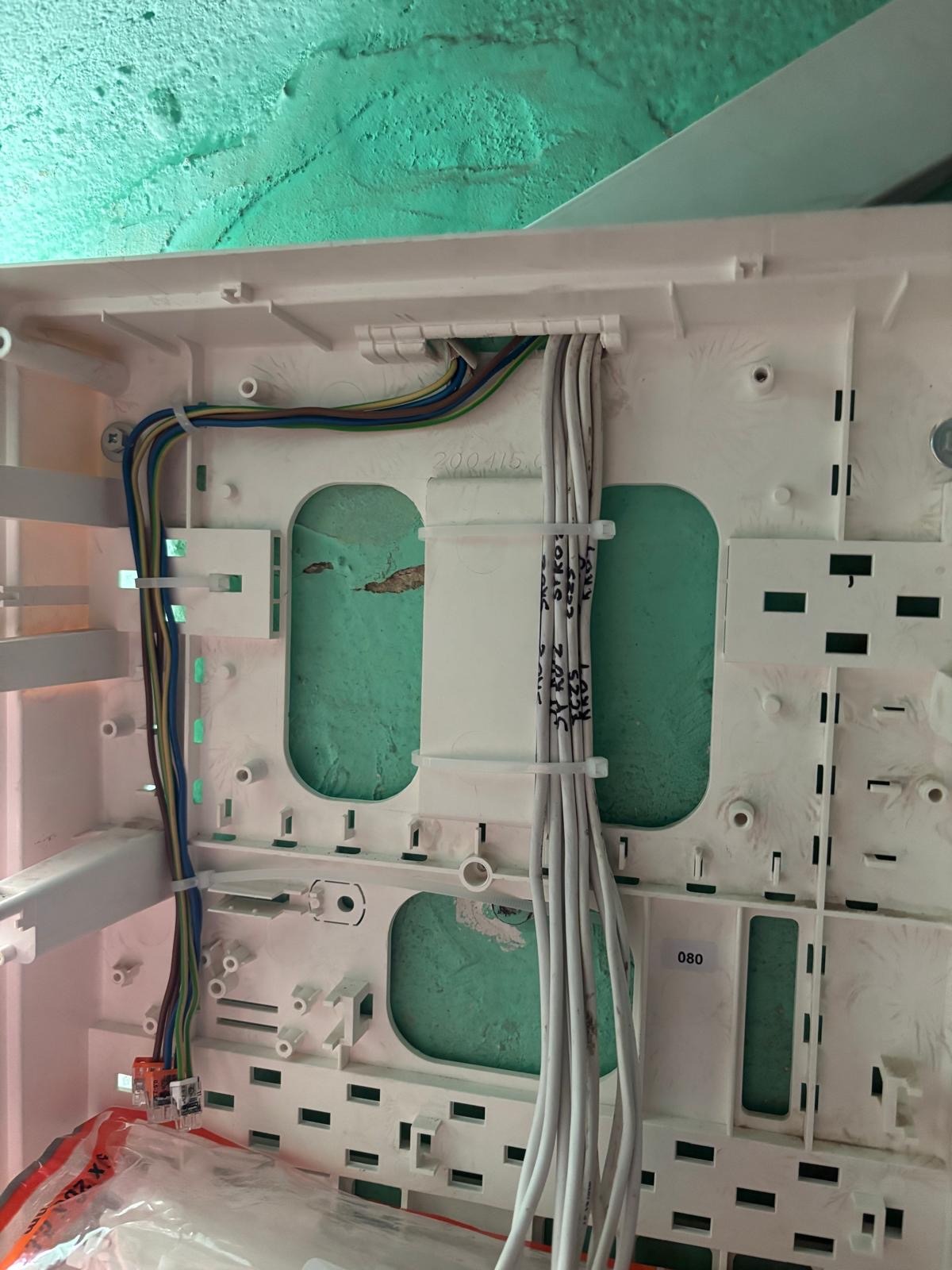 Wnętrze skrzynki elektrycznej z uporządkowanymi kablami, złączkami i opisami na przewodach, na tle zielonej ściany. Widoczne elementy montażowe i okablowanie.