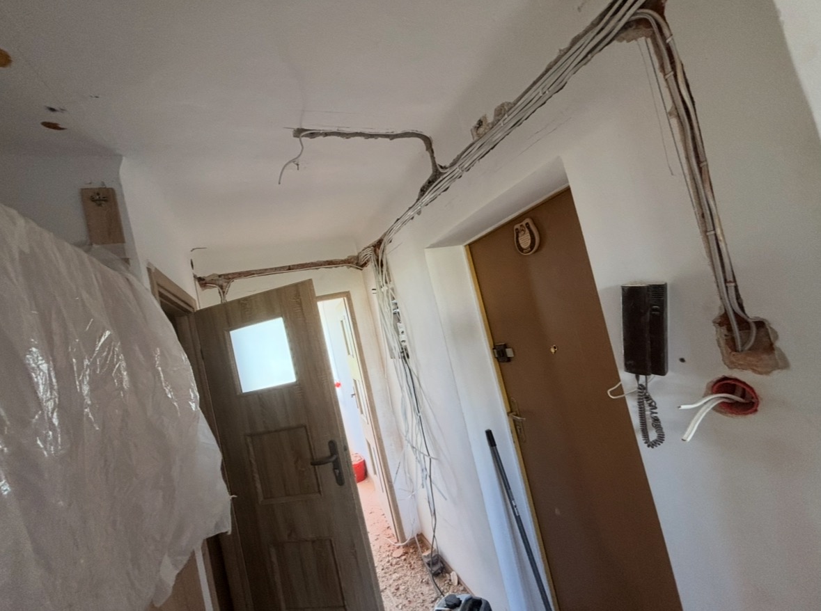Korytarz w trakcie remontu z widocznymi przewodami elektrycznymi ułożonymi wzdłuż sufitu i ścian, oraz zamontowany domofon przy brązowych drzwiach.