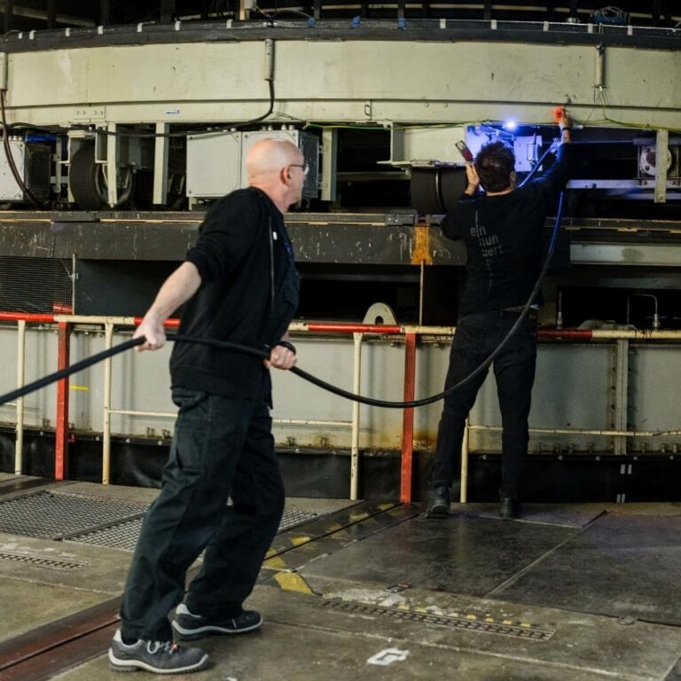 Dwóch mężczyzn pracuje przy dużej, okrągłej maszynie. Jeden ciągnie kabel, drugi oświetla obszar roboczy. Widoczne elementy elektryczne i metalowa konstrukcja.