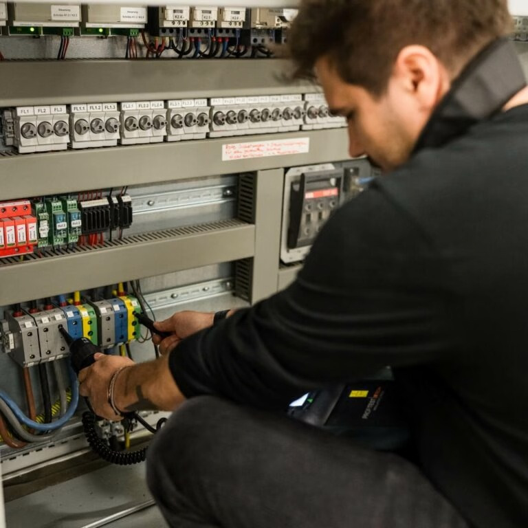 Elektryk w trakcie pracy: pomiary elektryczne w rozdzielnicy. Widoczne kable, bezpieczniki i miernik. Ujęcie z bliska, skupione na wykonywanych czynnościach.