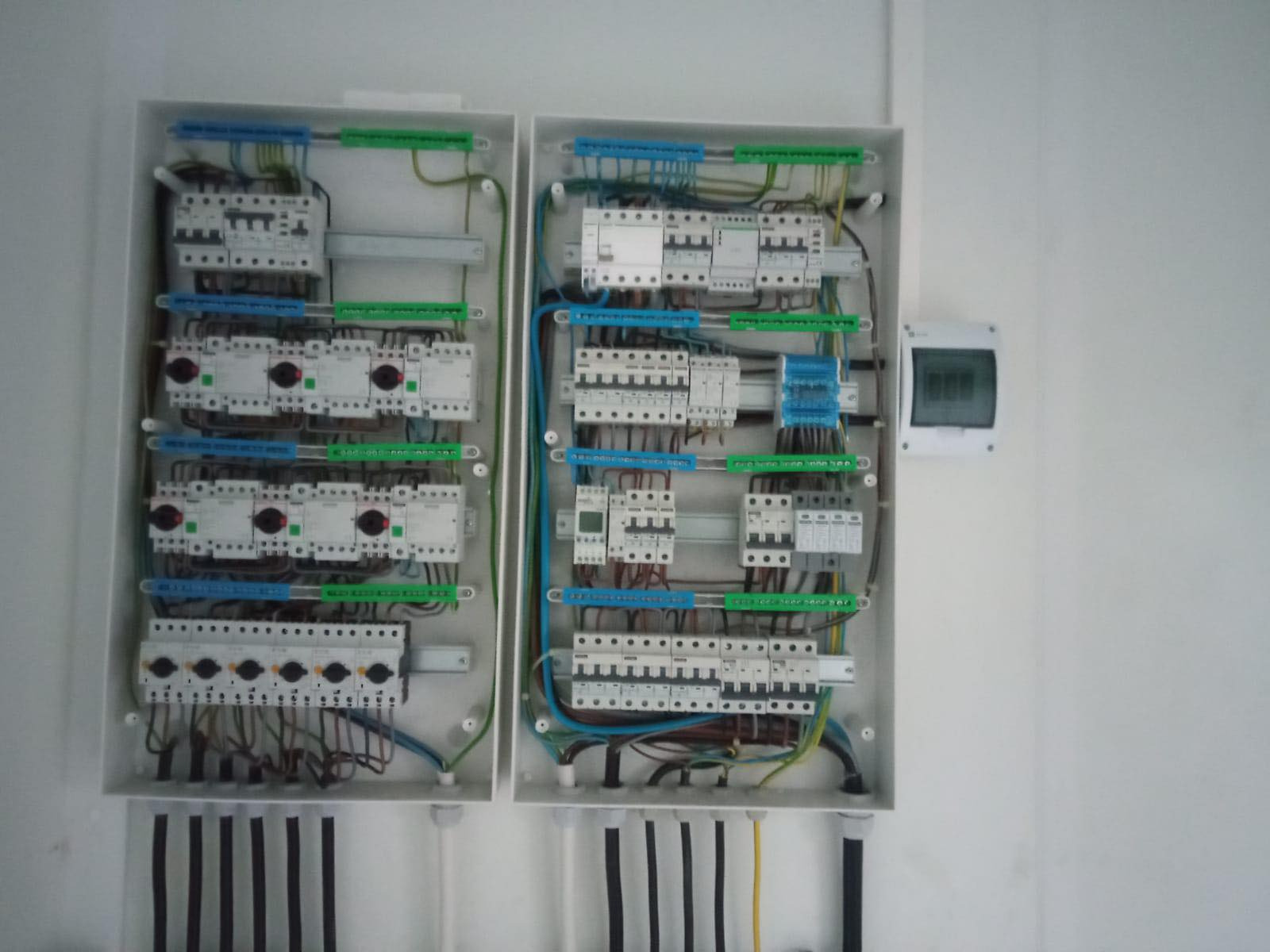 Dwa białe panele elektryczne z zainstalowanymi bezpiecznikami, przewodami i listwami, zamontowane na białej ścianie. Widoczne pionowe rury instalacyjne.