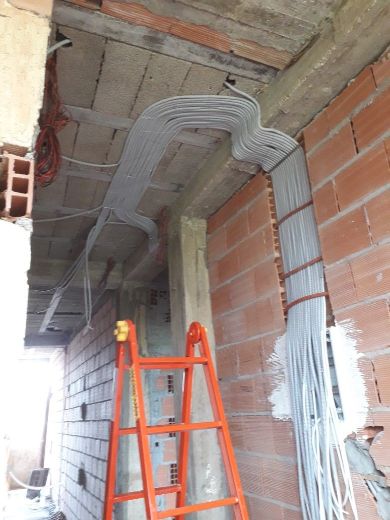 Szara instalacja elektryczna w peszlach ułożona wzdłuż ceglanej ściany w surowym stanie, widoczna pomarańczowa drabina.