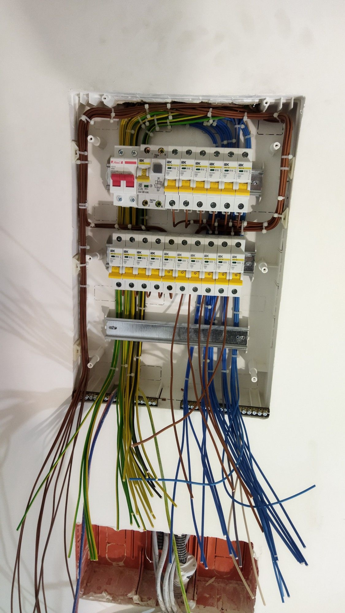 Rozdzielnica elektryczna z zabezpieczeniami IEK i widocznymi przewodami w kolorach brązowym, niebieskim i żółto-zielonym, zamontowana w białej obudowie w ścianie.