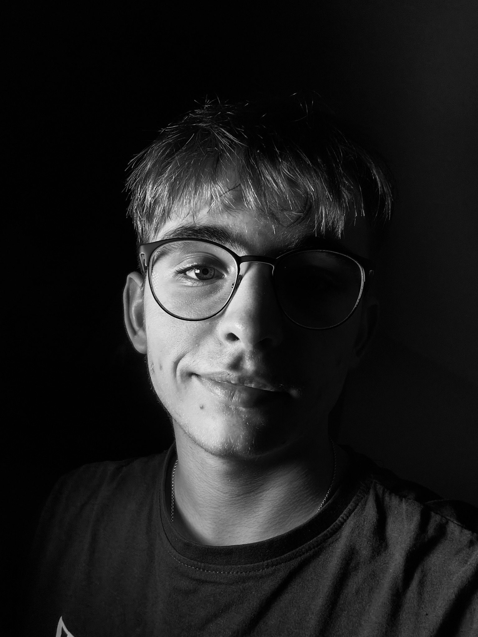 Czarno-biały portret młodego mężczyzny w okularach, oświetlonego z jednej strony, z ciemnym tłem. Widoczny uśmiech i detale twarzy.