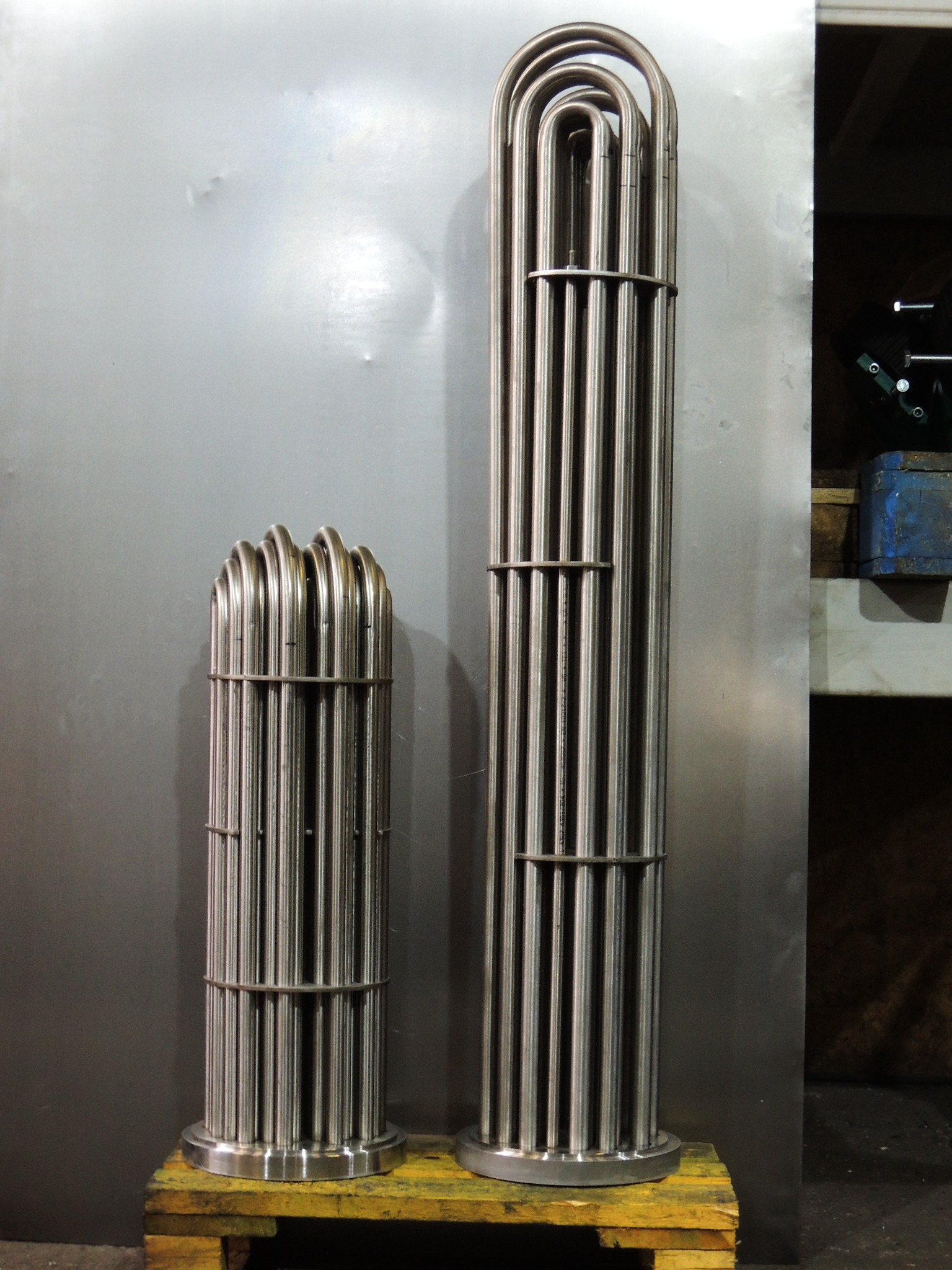 Dwa metalowe wymienniki ciepła na palecie, różnej wielkości. Większy, wysoki, z licznymi rurkami. Mniejszy, krępy, z zaokrąglonymi zakończeniami rurek.