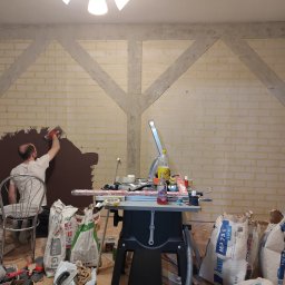 Piotr Płotka Uslugi Remontowo-Budowlane - Nakładanie brązowej gładzi na ścianę z imitacją cegły i drewnianych belek. W tle narzędzia i worki z zaprawą. Praca w toku, widok zza stanowiska pracy.