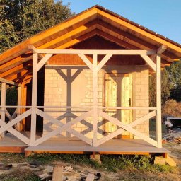 Piotr Płotka Uslugi Remontowo-Budowlane - Drewniana altana z jasną, krzyżową balustradą i ceglaną ścianą w tle, częściowo oświetlona słońcem, w otoczeniu zieleni. Widoczny etap budowy.