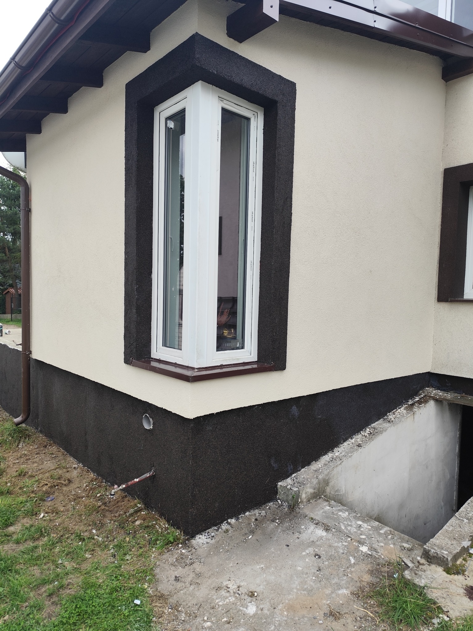 Wykończona elewacja domu z narożnym oknem, z kontrastowym, ciemnym cokołem i obramowaniem okna. Widoczne schody do piwnicy i fragment trawnika.