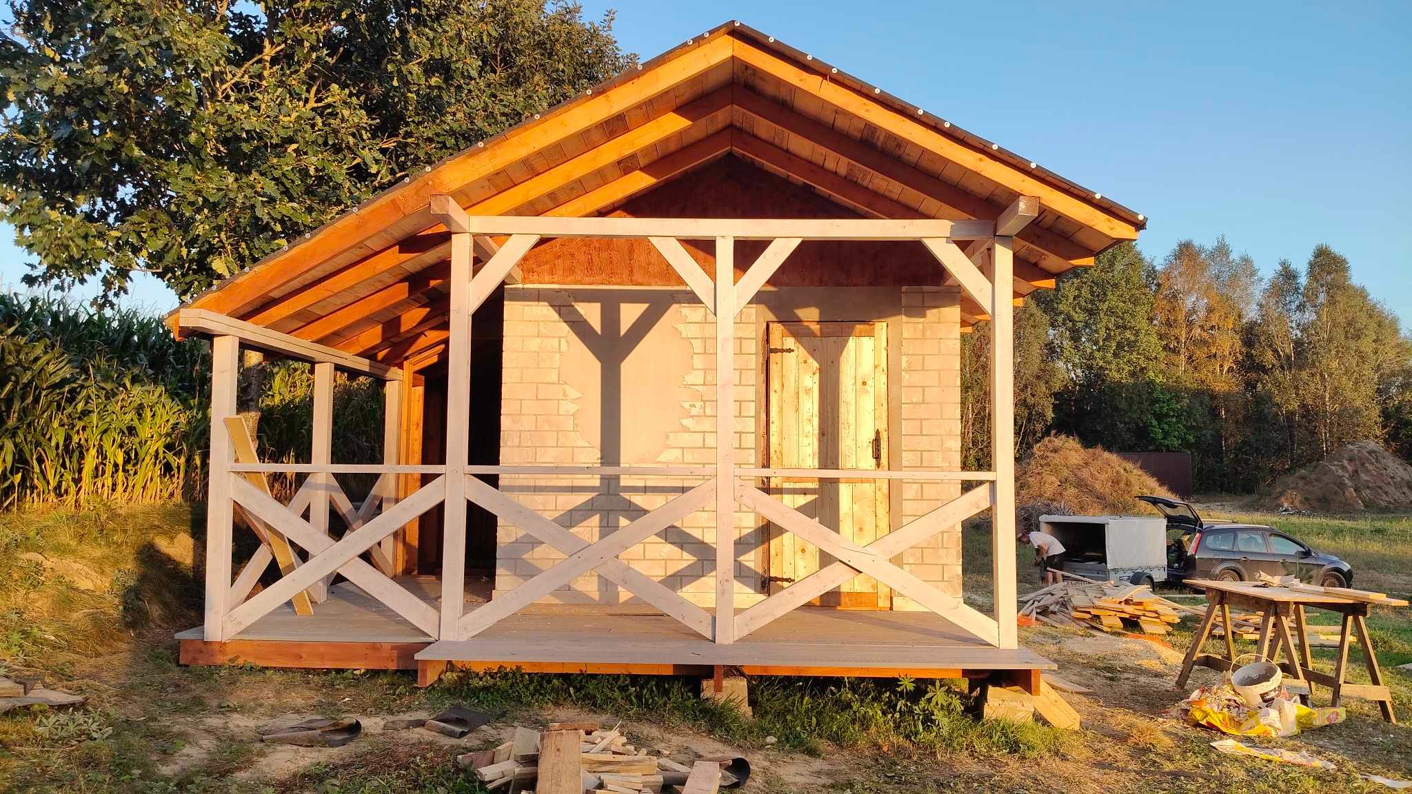Drewniana altana z jasną, krzyżową balustradą i ceglaną ścianą w tle, częściowo oświetlona słońcem, w otoczeniu zieleni. Widoczny etap budowy.