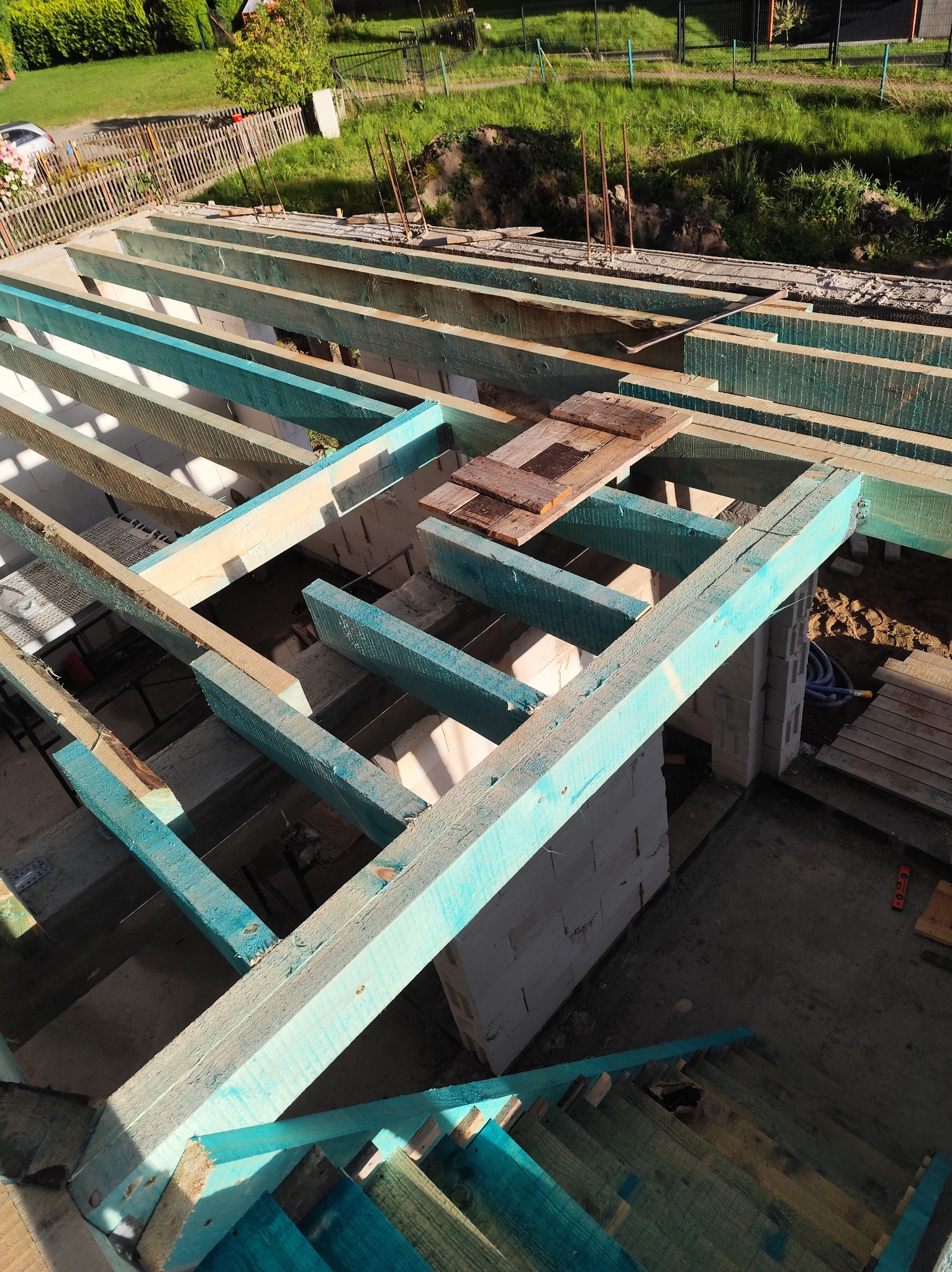 Konstrukcja drewniana z pomalowanymi na niebiesko belkami, prawdopodobnie tarasu, w trakcie budowy, widok z góry, z elementami muru i otaczającej zieleni.