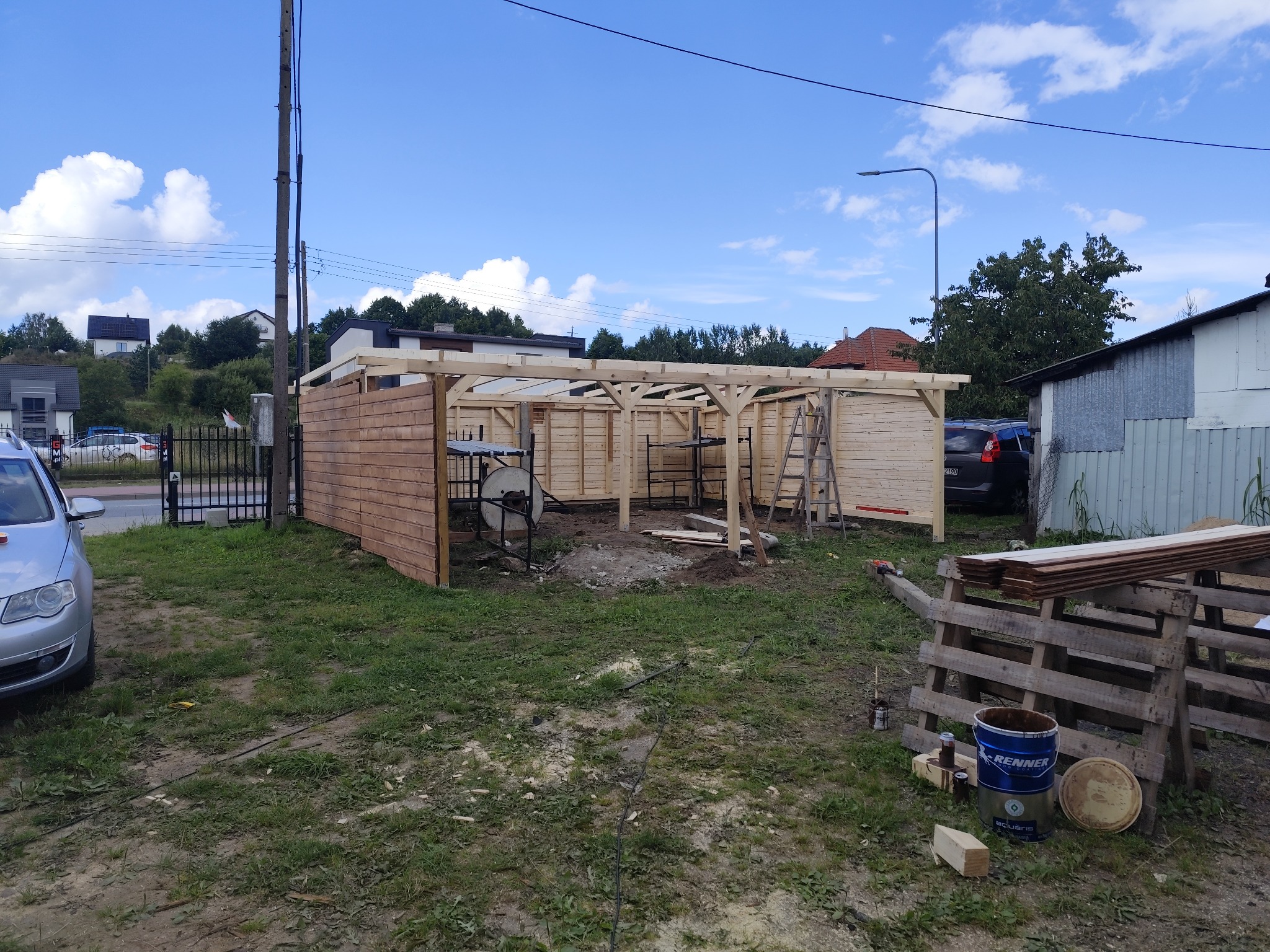 Wznoszenie drewnianej konstrukcji z desek na zewnątrz. Widoczne elementy szkieletu, częściowo obite ściany, drabina i puszki z impregnatem do drewna.