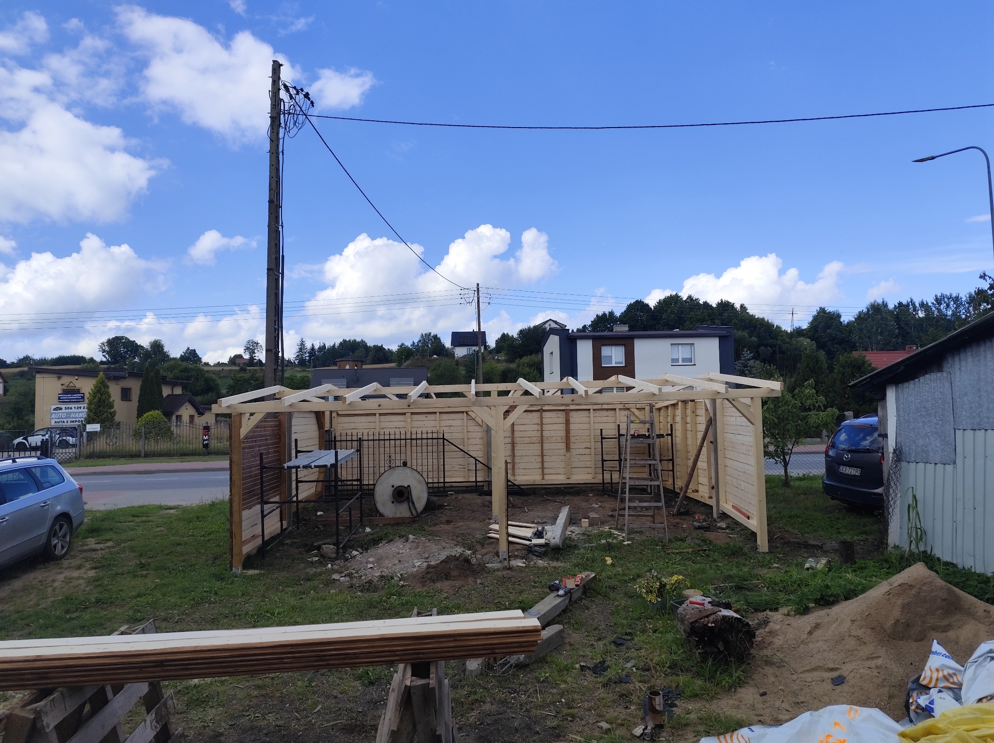 Drewniana konstrukcja w trakcie budowy, prawdopodobnie zadaszenie lub wiata, w ogrodzie, z widocznymi elementami konstrukcyjnymi i narzędziami.