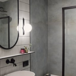 ALZA STUDIO Alicja Zapolna - Nowoczesna łazienka z owalnym lustrem, umywalką nablatową i płytkami w pionowe paski. Minimalistyczny design, szare ściany, czarne akcenty i drewniane szafki.