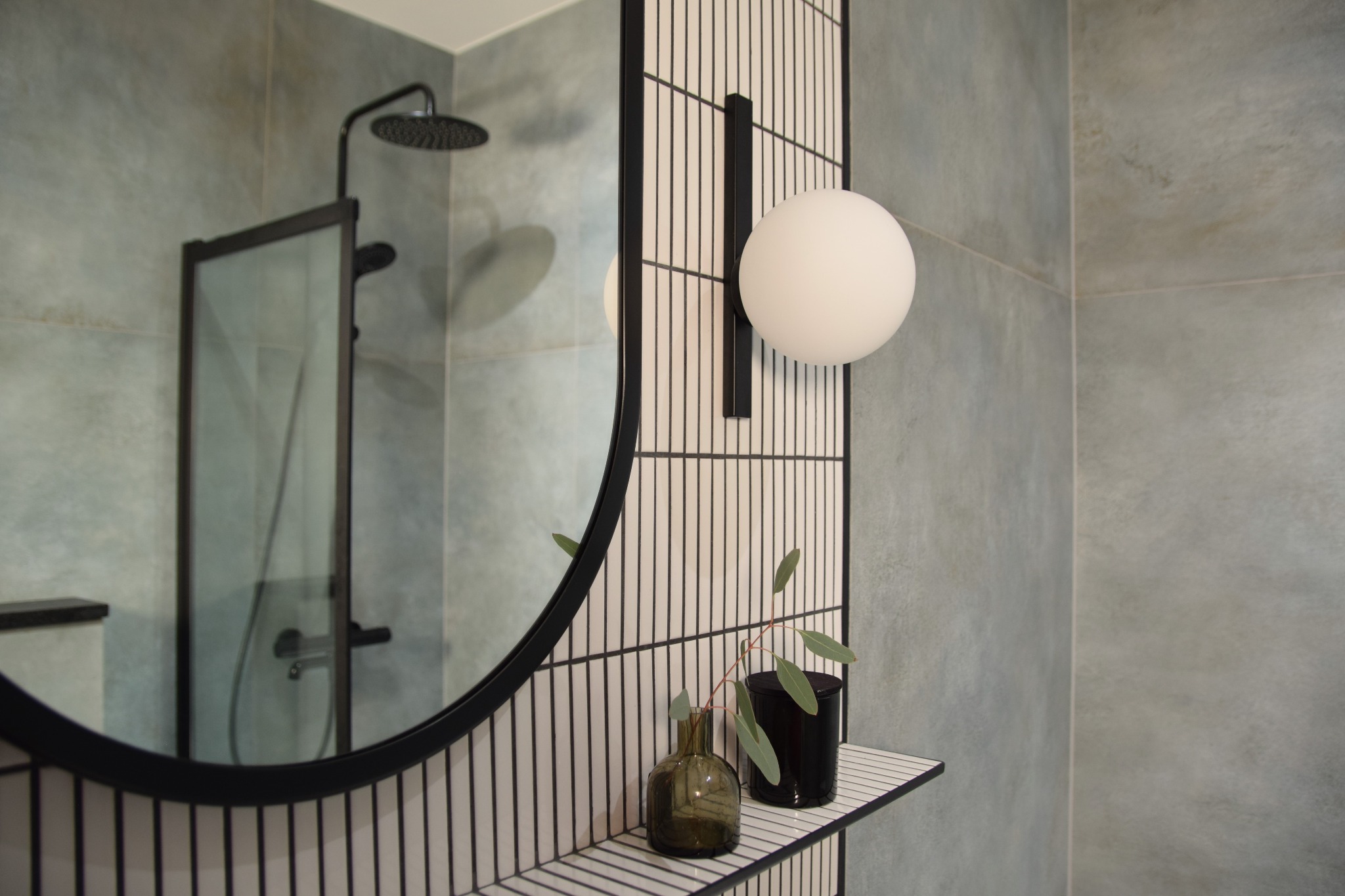 Nowoczesna łazienka z okrągłym lustrem w czarnej ramie, oświetleniem kulistym i minimalistycznymi akcentami roślinnymi na półce z płytek.