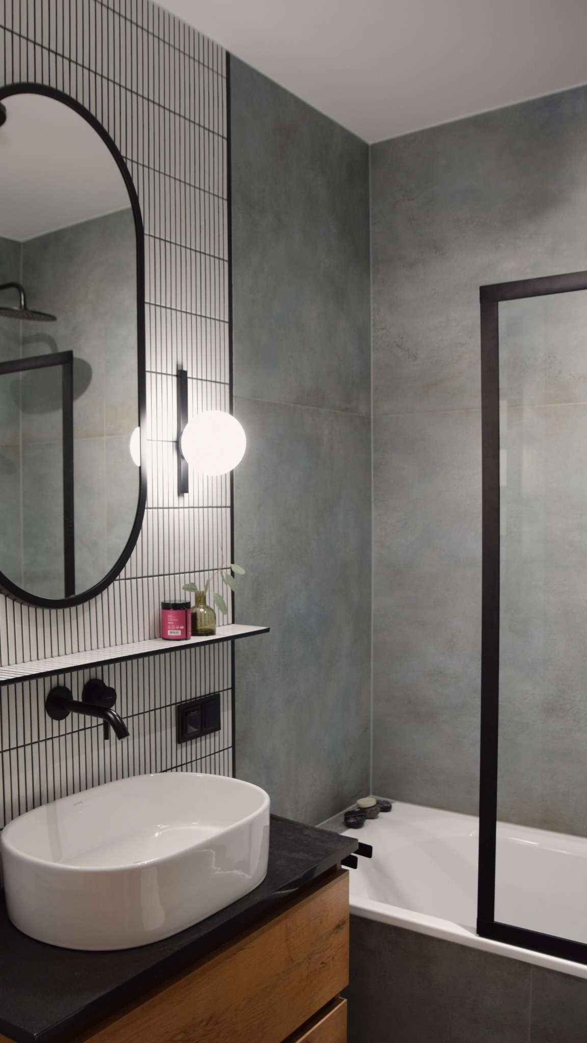Nowoczesna łazienka z owalnym lustrem, umywalką nablatową i płytkami w pionowe paski. Minimalistyczny design, szare ściany, czarne akcenty i drewniane szafki.