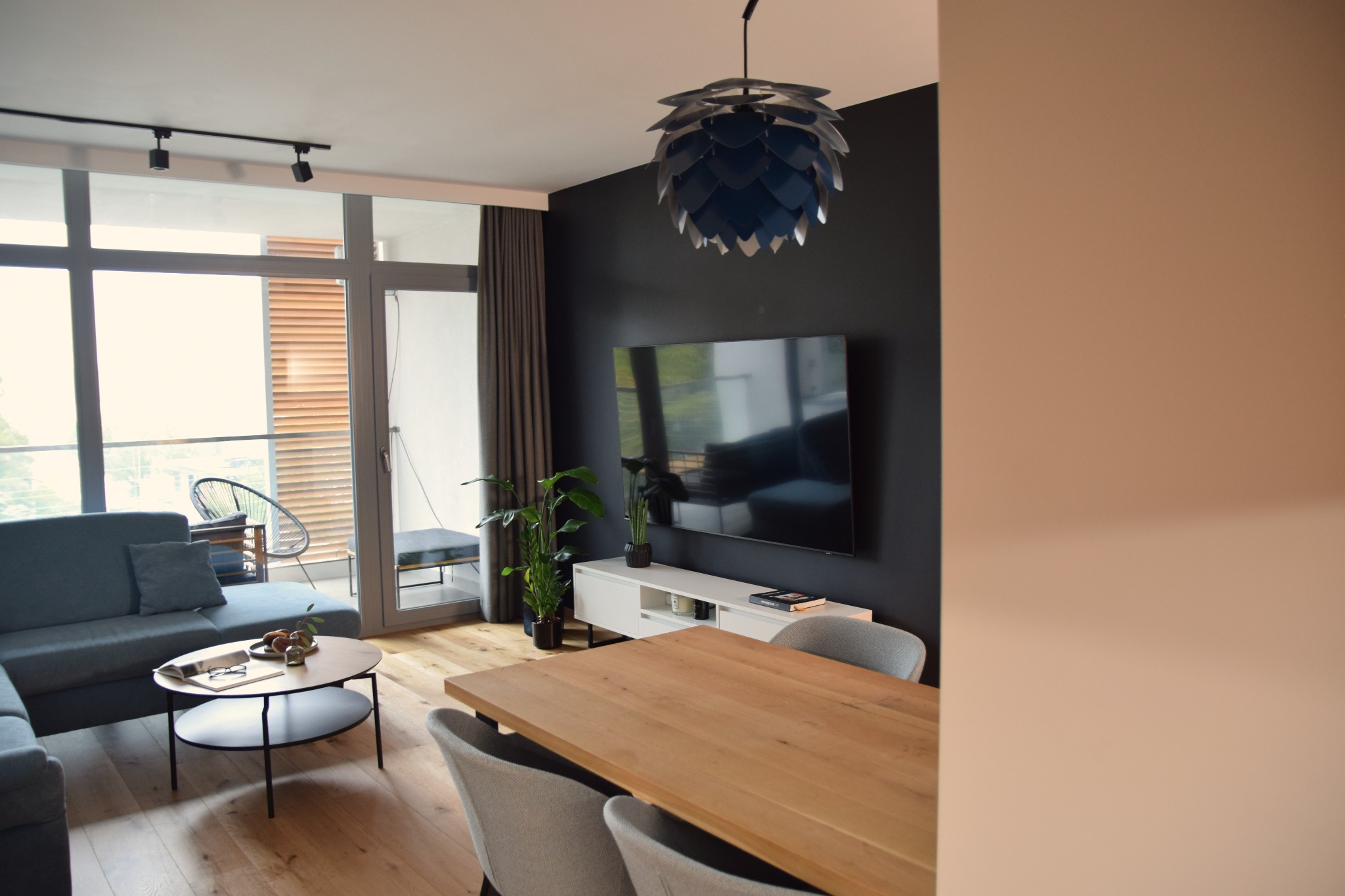 Nowoczesny salon z drewnianym stołem, szarą sofą i balkonem. Ciemna ściana za telewizorem, designerska lampa. Minimalistyczny i elegancki wystrój.