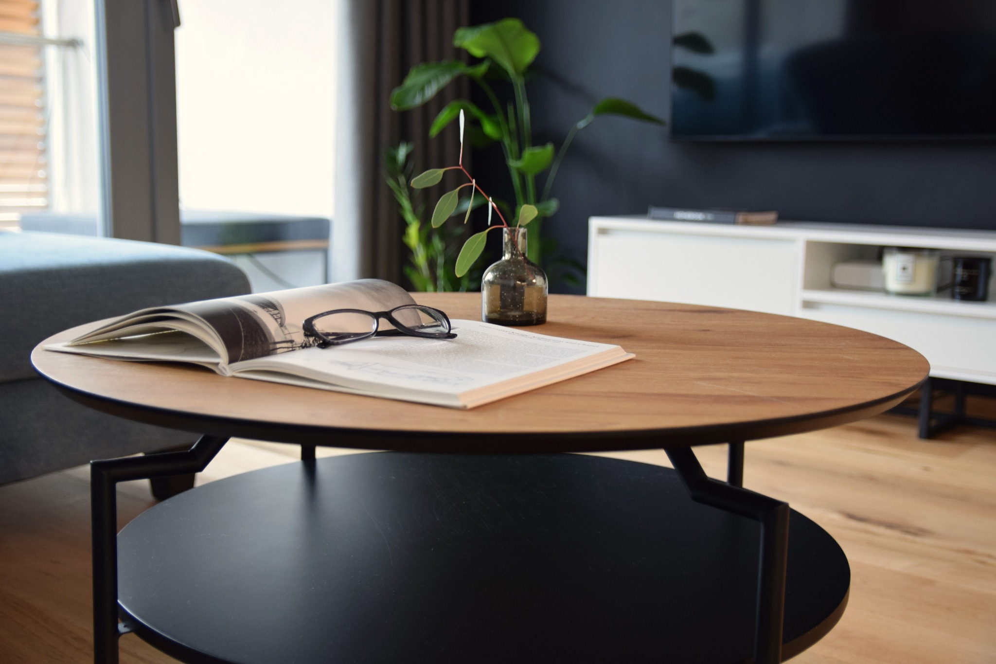 Stylowy stolik kawowy z drewnianym blatem i czarną podstawą. Na nim otwarta książka z okularami i wazonik z gałązką. W tle minimalistyczny salon z szarą ścianą.