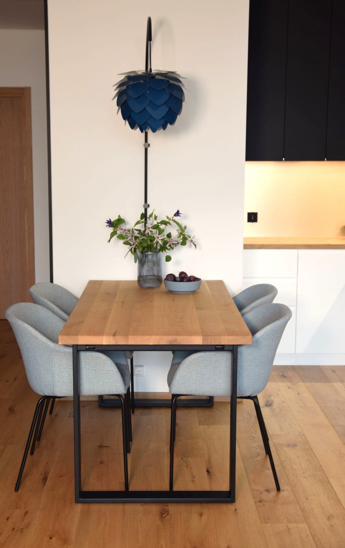 Nowoczesna jadalnia: stół z drewna, szare krzesła, designerska lampa w kształcie szyszki. Wazon z kwiatami i miska ze śliwkami na blacie. Minimalistyczny wystrój.