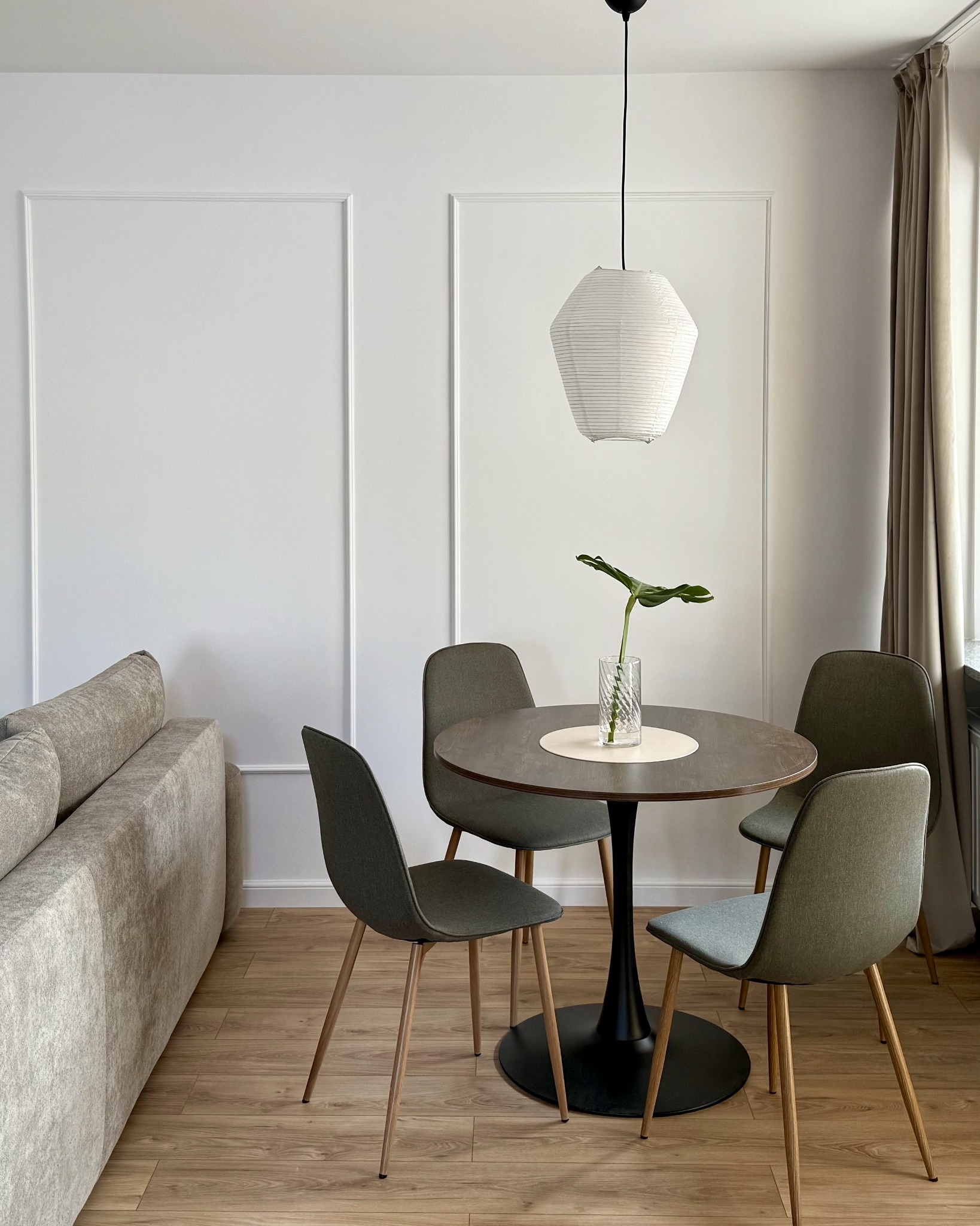 Jasne wnętrze: okrągły stół z czterema krzesłami, minimalistyczna lampa, sofa i dekoracyjne panele ścienne. Parkiet na podłodze, stonowana kolorystyka.