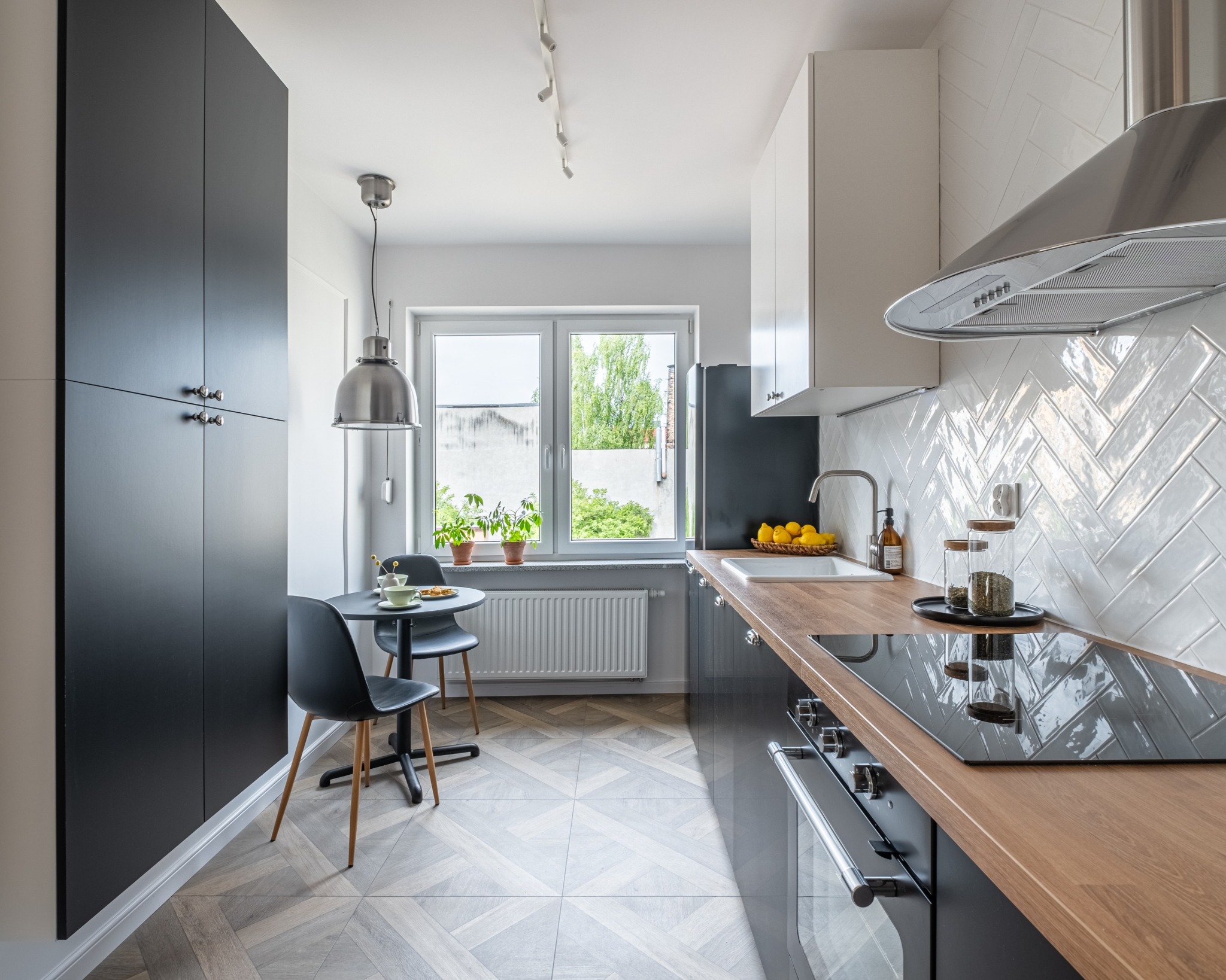 Nowoczesna kuchnia z czarnymi szafkami, drewnianym blatem i białymi płytkami ułożonymi w jodełkę. Stół z krzesłami przy oknie, nad nim lampa. Jasne, przestronne wnętrze.