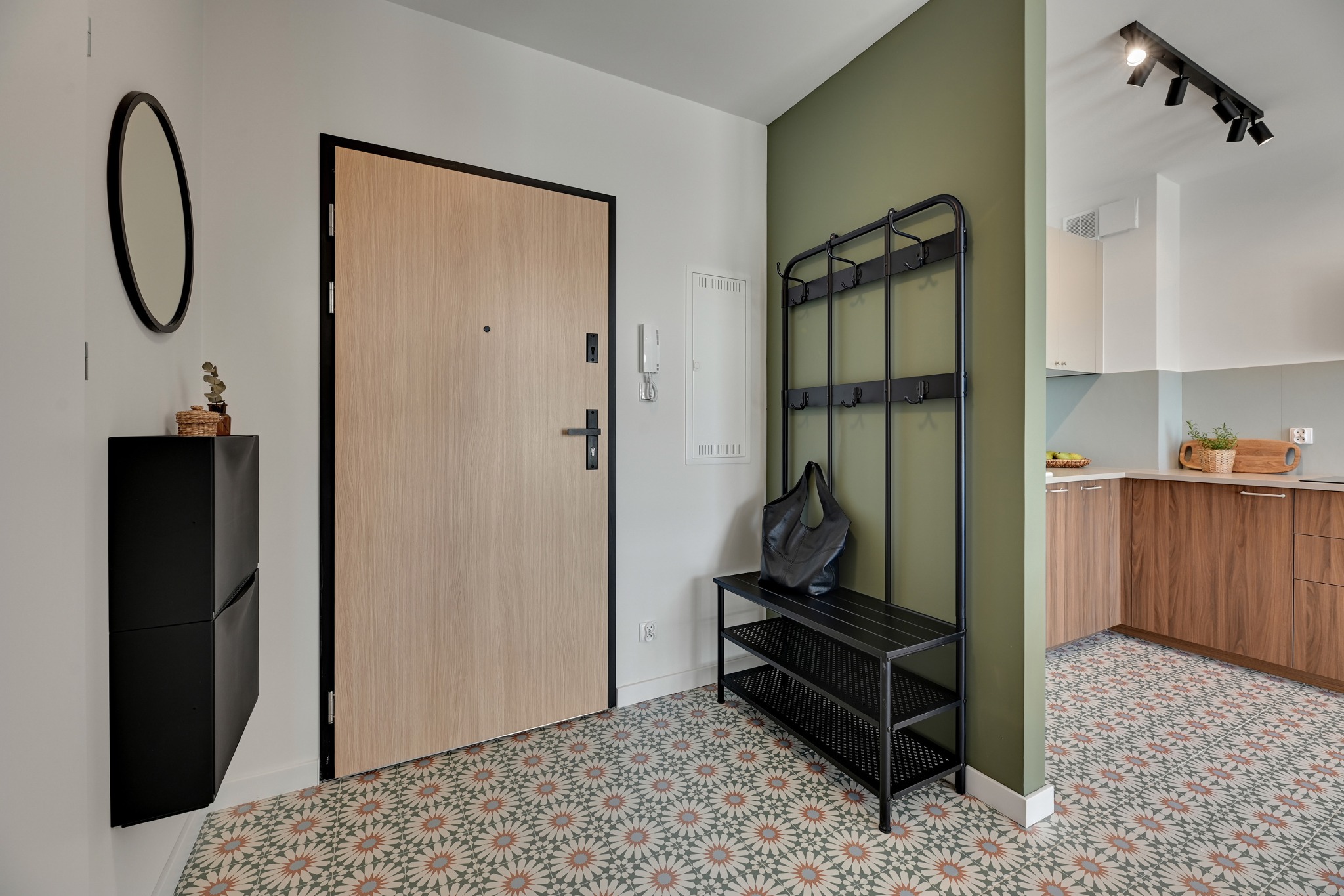 Nowoczesny przedpokój z drewnianymi drzwiami, czarną szafką na buty i wieszakiem. W tle widoczna kuchnia z drewnianymi szafkami i wzorzysta podłoga.
