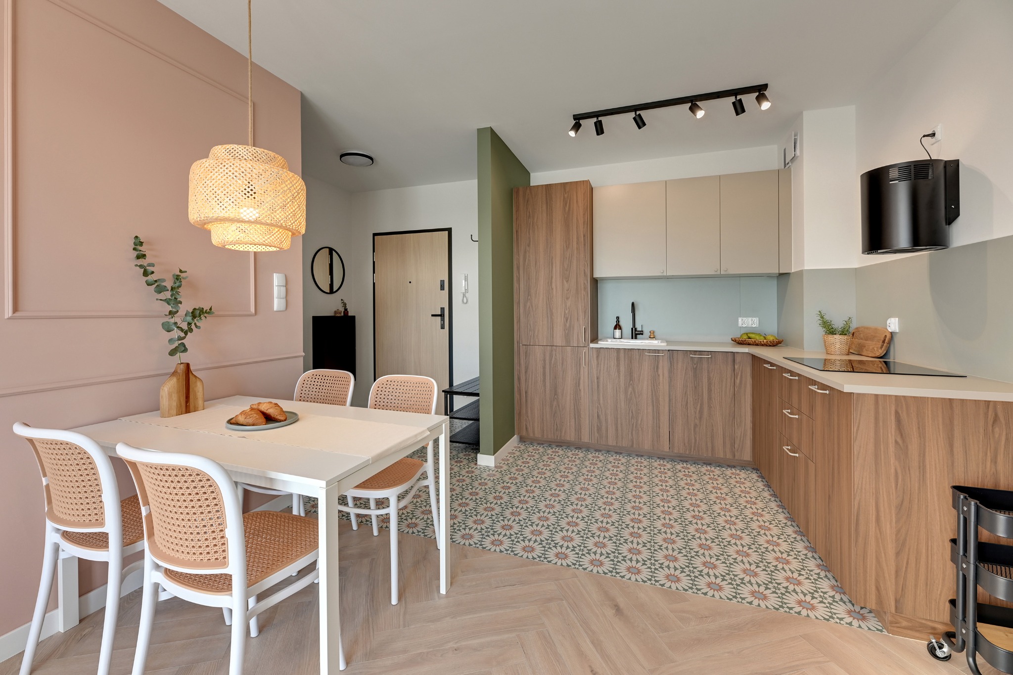 Jasne, nowoczesne wnętrze: stół z wiklinowymi krzesłami, lampa z abażurem, kuchnia z drewnianymi frontami i wzorzystą podłogą. Pastelowe kolory ścian.