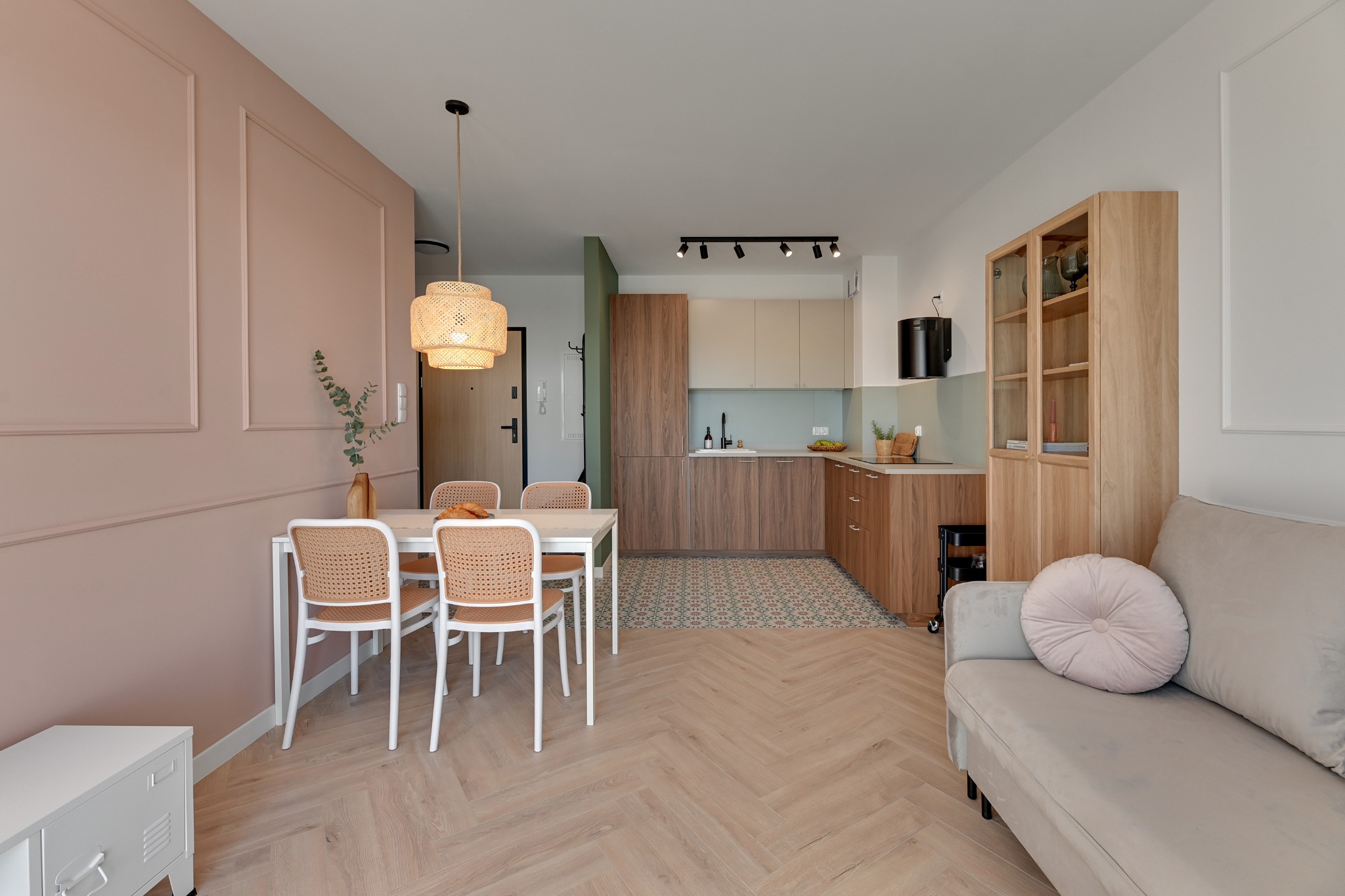 Jasna, przestronna kuchnia z jadalnią: drewniane szafki, biały stół z rattanowymi krzesłami, parkiet w jodełkę, różowa ściana z dekoracyjnymi listwami.