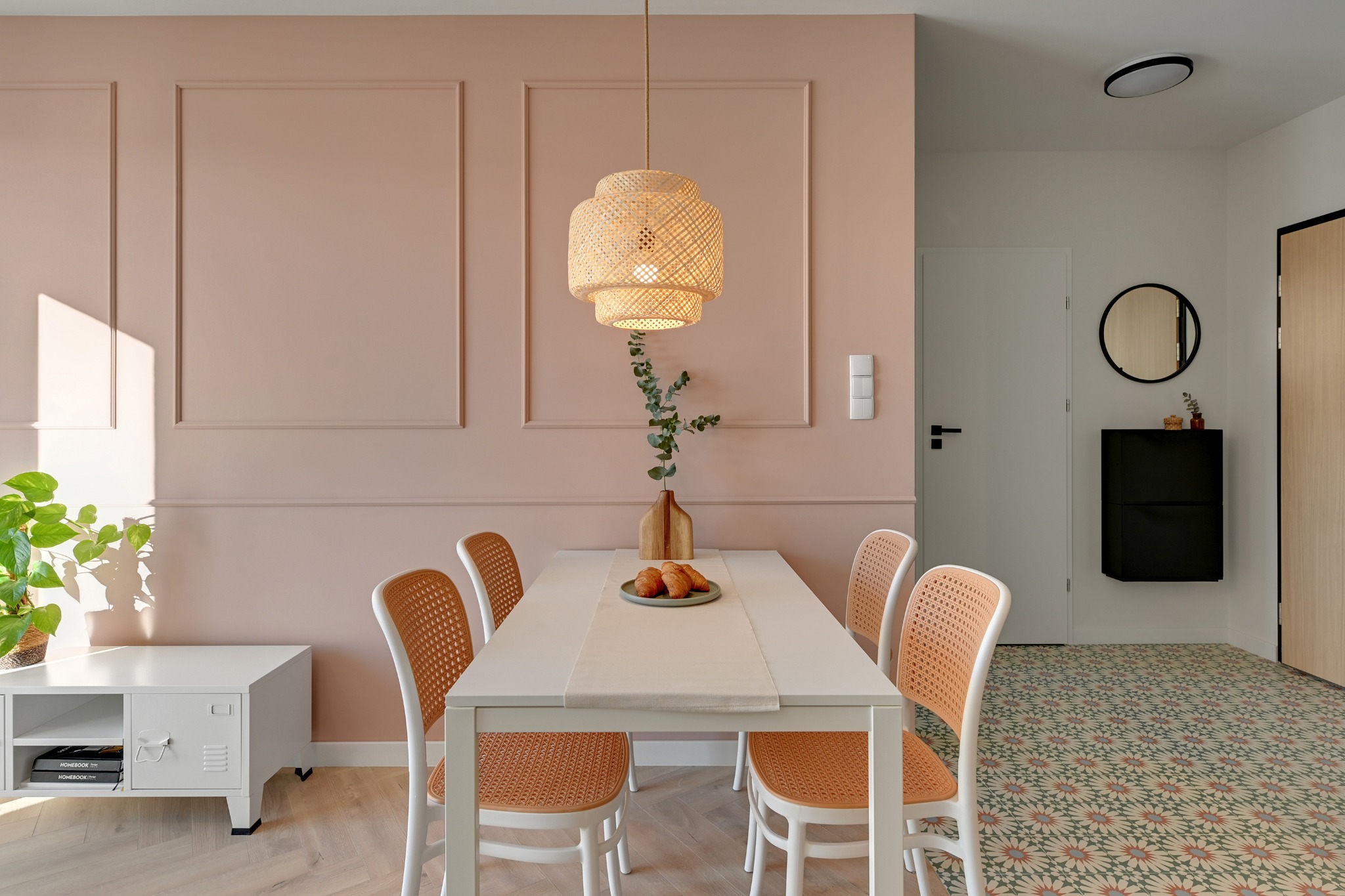 Jasna jadalnia z różową ścianą, białym stołem i rattanowymi krzesłami. Nad stołem wisi lampa z abażurem z rattanu. W tle widoczny przedpokój z okrągłym lustrem.