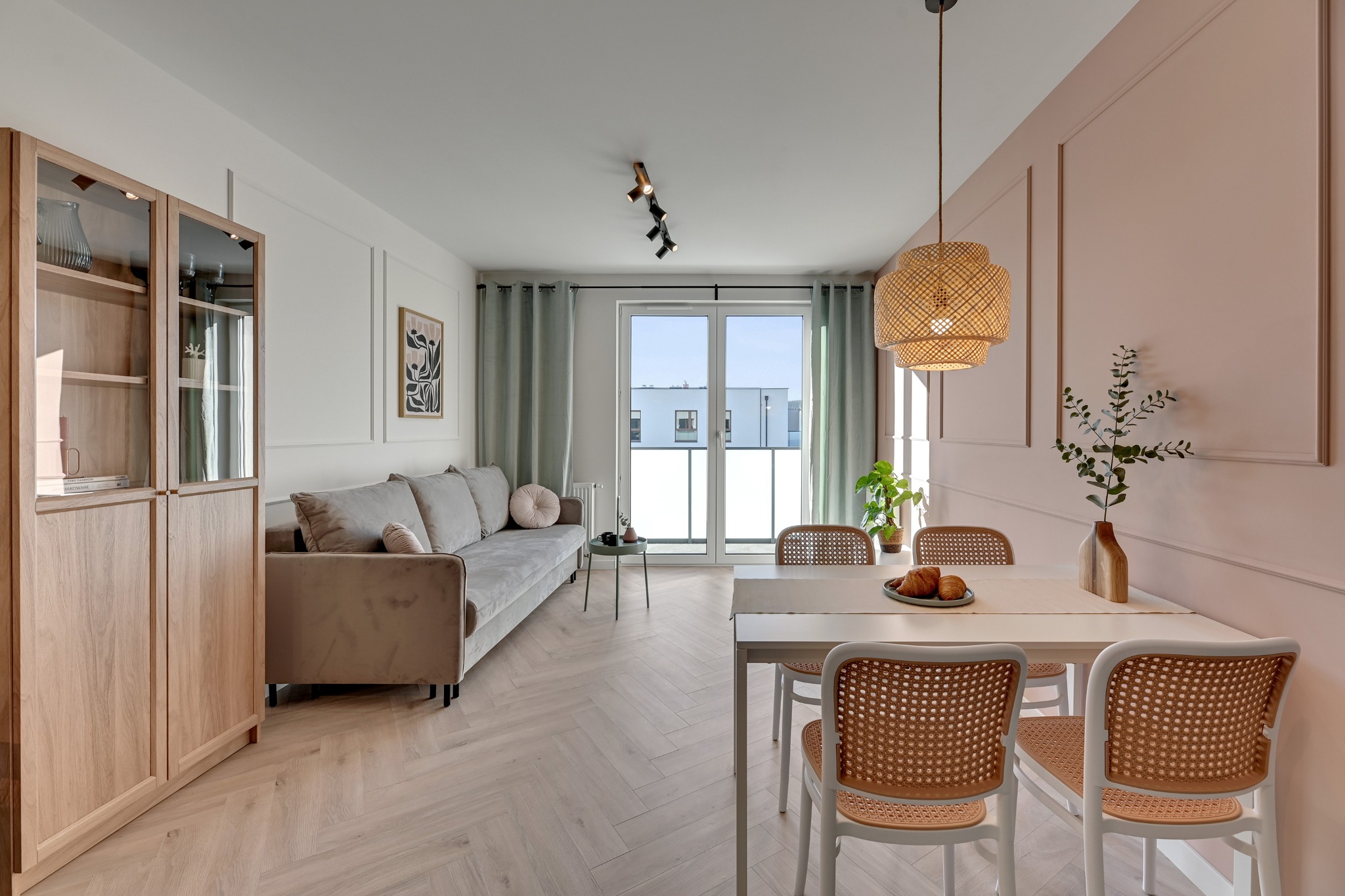 Jasne wnętrze salonu z jadalnią: sofa, stół z rattanowymi krzesłami, drewniana witryna i dekoracyjna lampa wisząca. Pastelowa kolorystyka, parkiet w jodełkę.