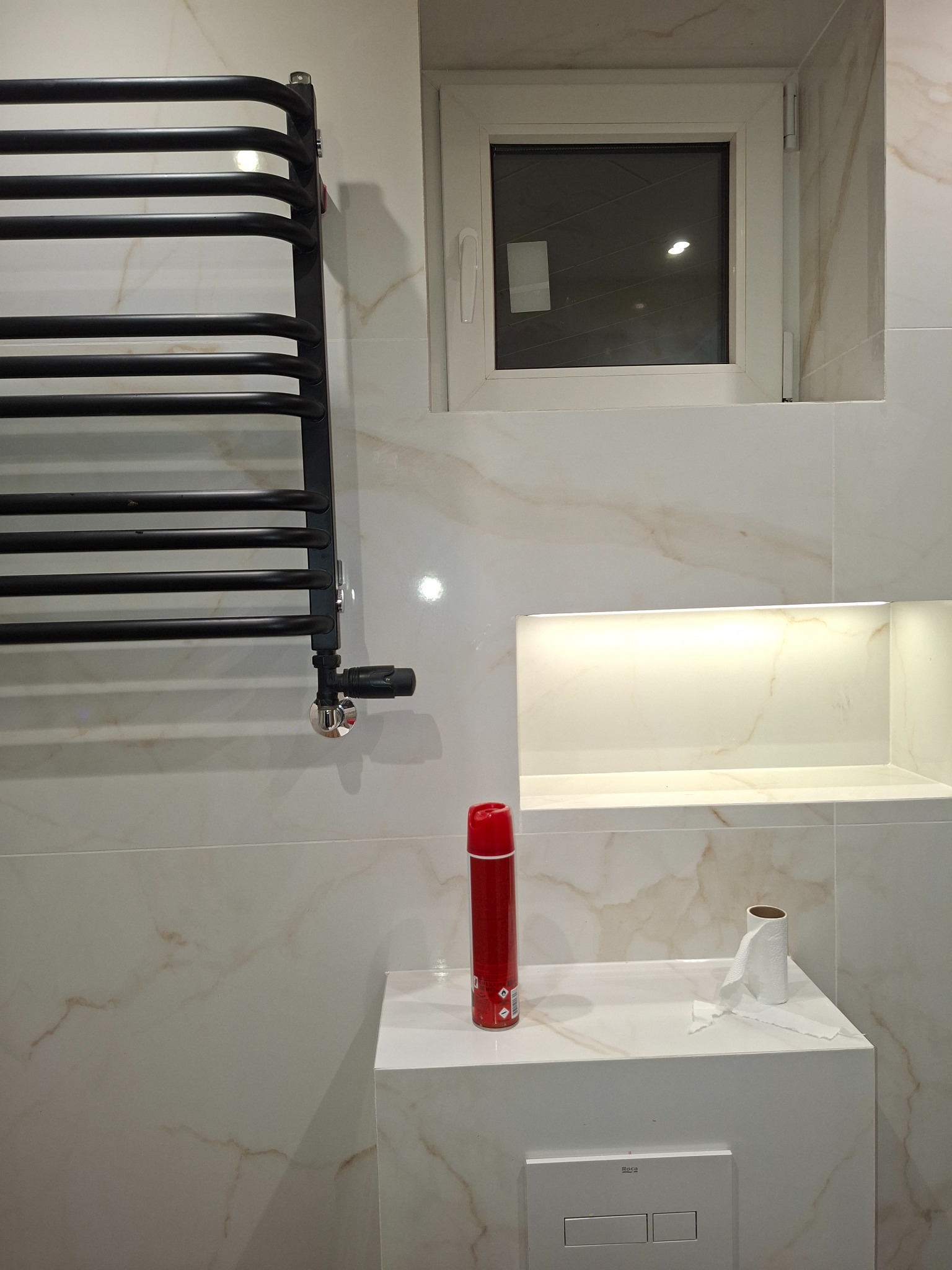 Wykończona łazienka z marmurowymi płytkami, czarnym grzejnikiem drabinkowym, podświetlaną wnęką i podtynkową toaletą z czerwonym odświeżaczem powietrza.