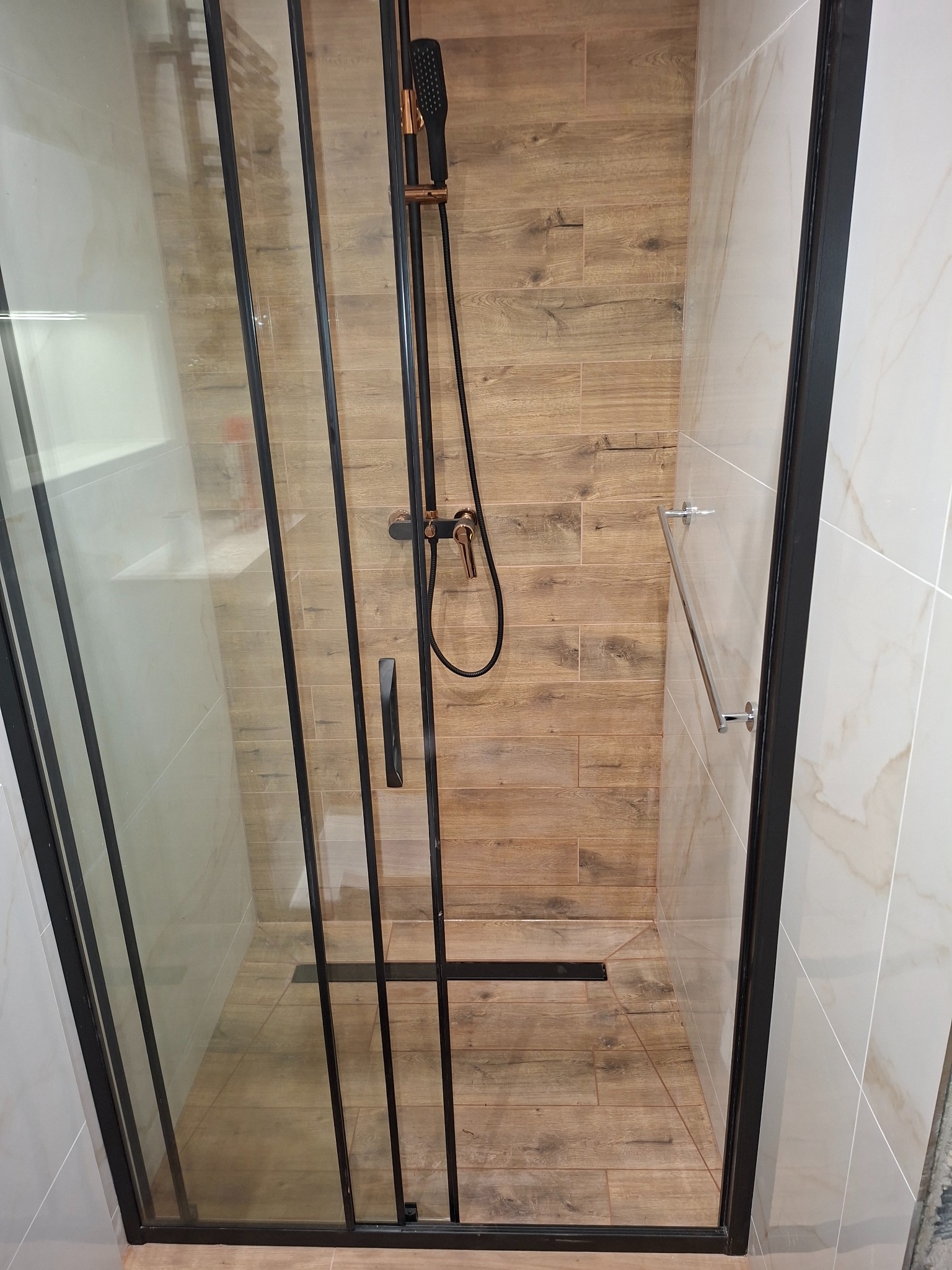 Nowoczesna kabina prysznicowa z czarną ramą i armaturą w kolorze miedzi, wykończona płytkami imitującymi drewno i marmur, z odpływem liniowym w podłodze.