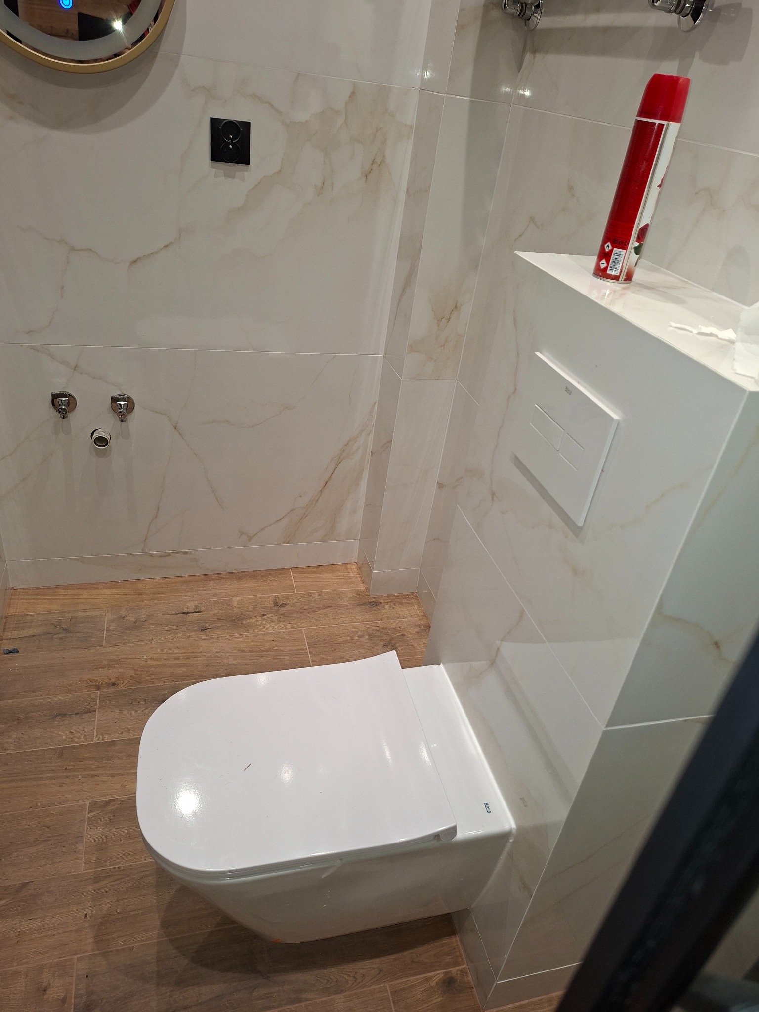 Nowoczesna toaleta podwieszana w wykończonej łazience z marmurowymi płytkami i drewnopodobną podłogą. Widoczny dozownik i czarny kontakt.