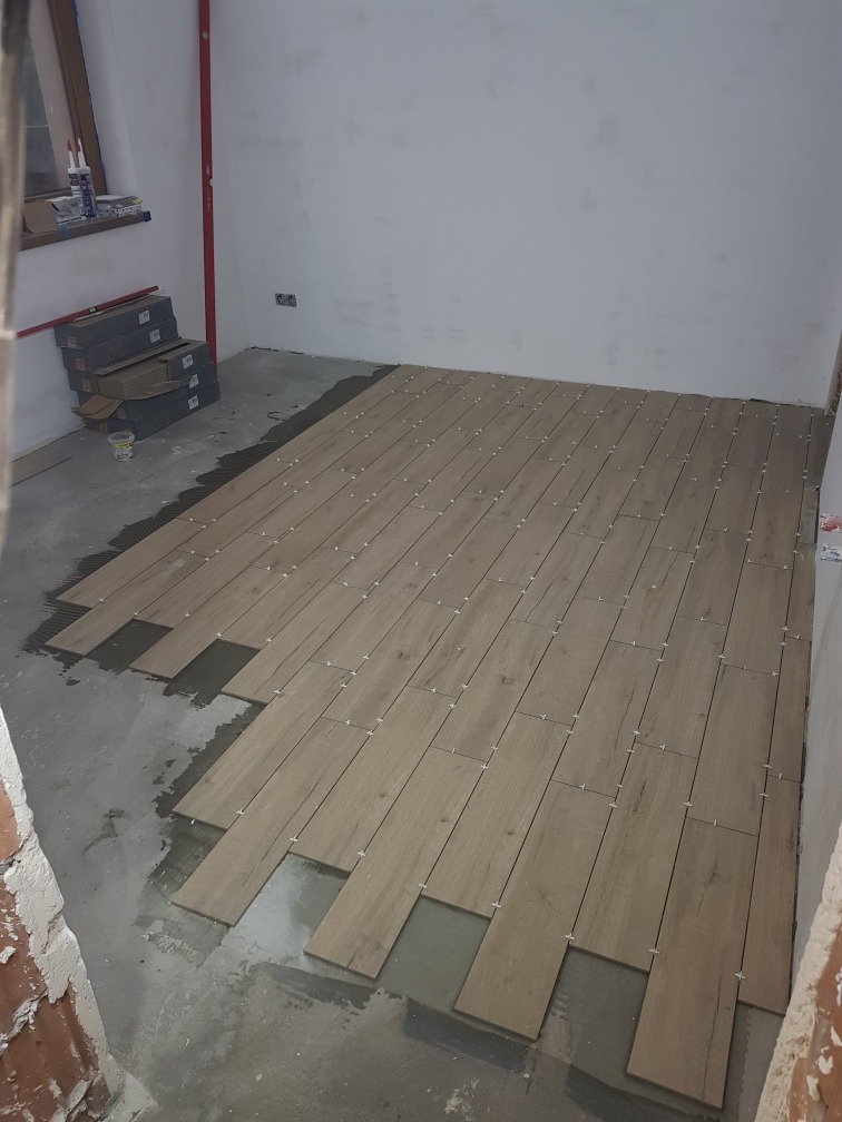 Układanie paneli podłogowych imitujących drewno w jasnym pomieszczeniu. Widoczne płytki, klej i krzyżyki dystansowe. Częściowo ułożona podłoga, surowe ściany.