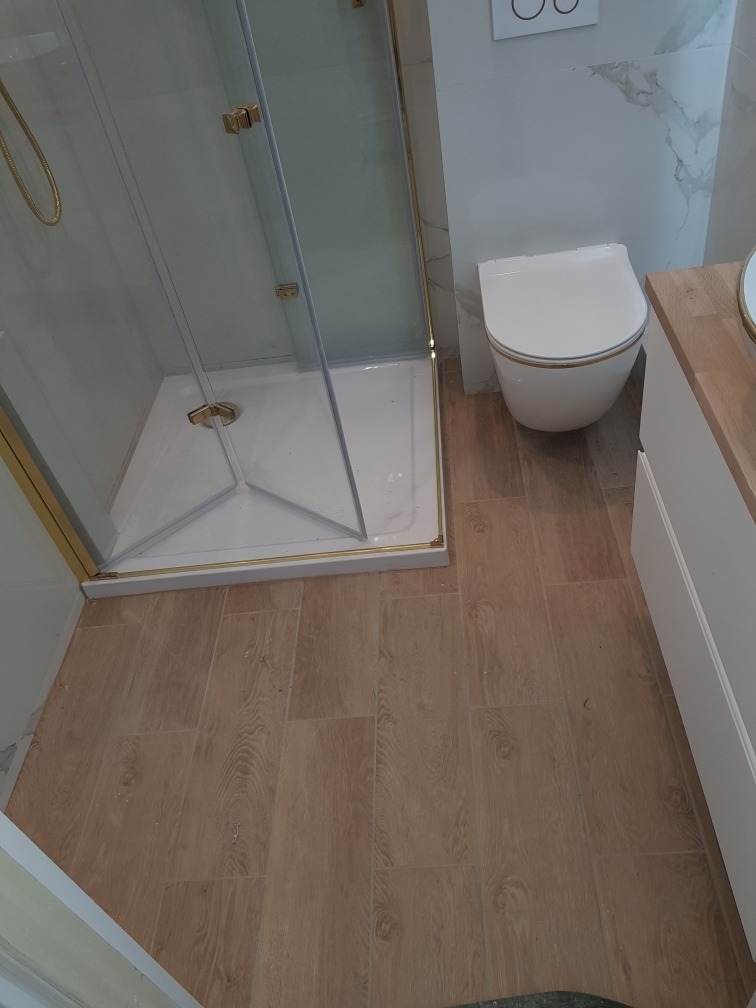 Nowoczesna łazienka po remoncie: kabina prysznicowa ze złotymi detalami, podwieszana toaleta i drewnopodobne płytki podłogowe. Jasne, minimalistyczne wykończenie.