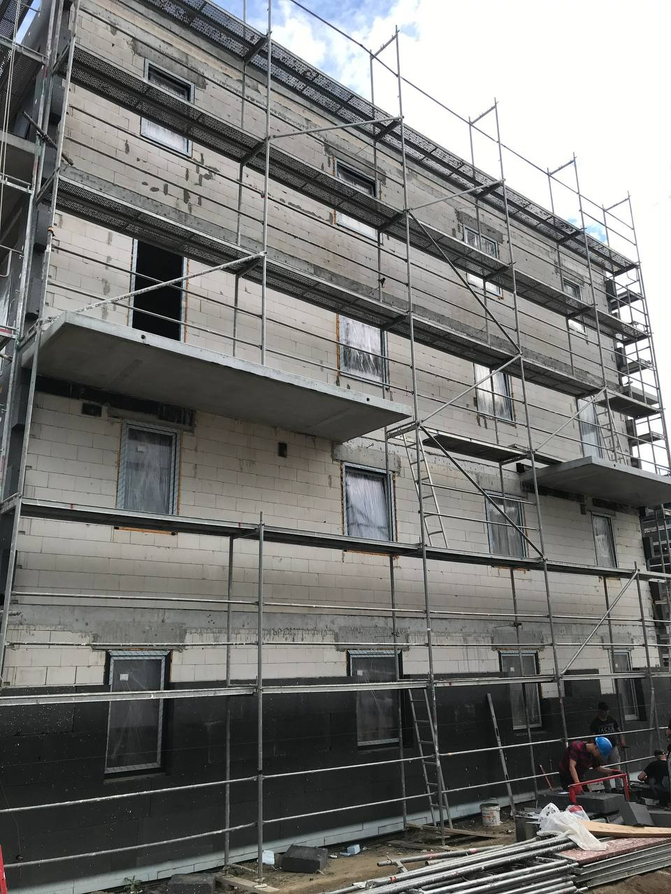 Budynek w trakcie ocieplania, widoczne rusztowania i pracownicy. Dolna część elewacji w kolorze czarnym, górna w jasnym szarym. Okna zabezpieczone folią.