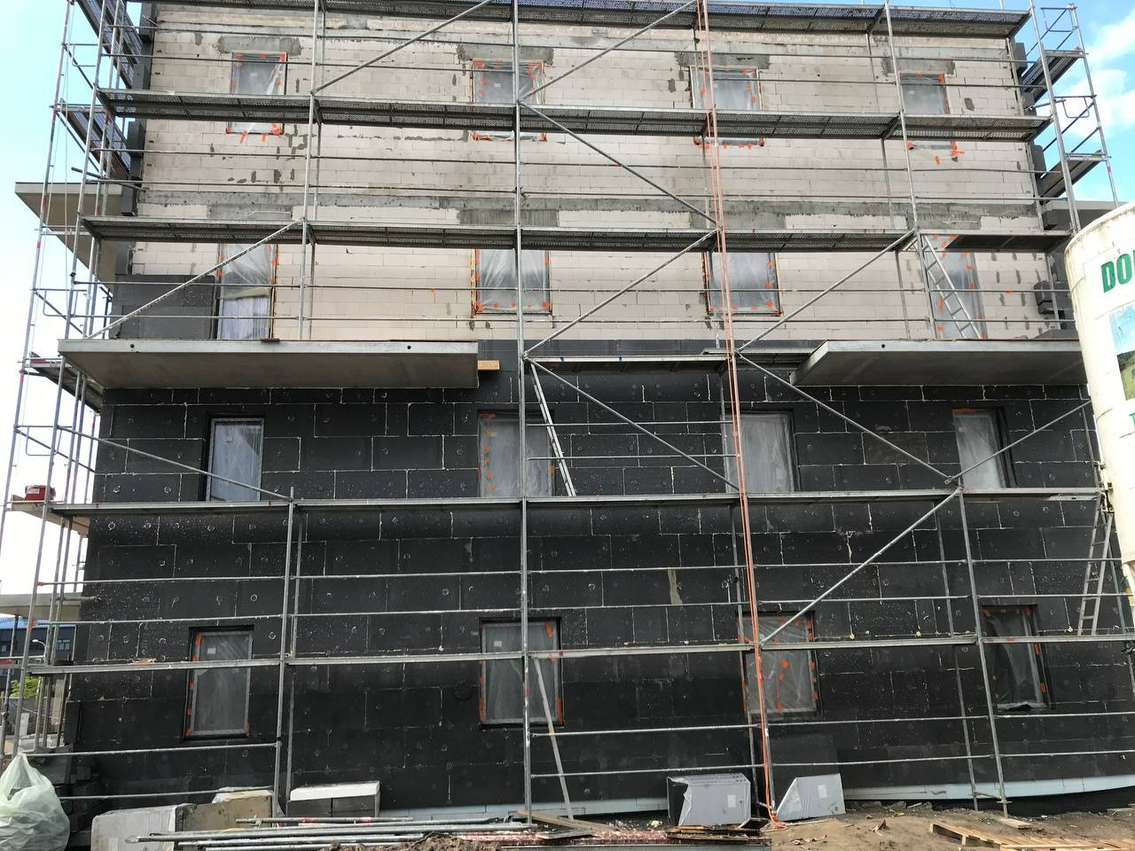 Budynek w trakcie ocieplania styropianem grafitowym, zabezpieczone okna folią, widoczne rusztowania w Warszawie. Prace elewacyjne na różnych etapach.